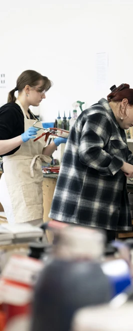 Two people working at a printmaking studio workbench, using tools and materials, with art supplies and equipment visible in the foreground and background Two people working at a printmaking studio workbench, using tools and materials, with art supplies and equipment visible in the foreground and background